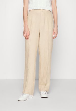 Persoon draagt hoge beige wijde broeken gecombineerd met witte sneakers en een wit T-shirt met korte mouwen tegen een effen achtergrond.