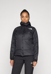 Žena u crnoj podstavljenoj jakni i hlačama The North Face, stoji naslonjena na jednostavnu svijetlosivu pozadinu, okrenuta prema naprijed.