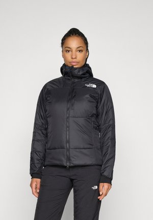 Woman wearing black The North Face padded jacket and pants, standing against a plain light gray background, facing forward.