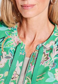 Groene bloemen-jas met roze, blauwe en gele patronen. Voorzien van een ritsluiting en een trekkoordkap. Gladde stoftextuur.