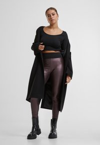 Palton lung negru, top negru mulat și colanți lucioși burgundy. Bocanci negri masivi. Modelul stă în fața unui fundal alb simplu.