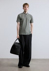 Γκρι πόλο πουκάμισο με λευκή επένδυση, φορεμένο με φαρδιά μαύρη παντελόνια. Το μοντέλο κρατά μια μαύρη δερμάτινη τσάντα duffel. Απλός, μοντέρνος σχεδιασμός.