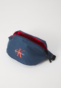 Kék szövet övtáska piros belsővel, elülső cipzáras zsebbel, és narancssárga-fehér Calvin Klein logóval. Állítható pánt tartozik hozzá.