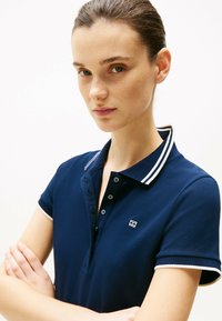 Granatowa koszulka polo z krótkim rękawem, biało-szary kołnierzyk w paski, zapięcie na sześć guzików oraz małe haftowane logo na piersi.