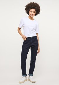 Tumši zilie džinsi ar šauru piegriezumu, piecu kabatu dizainu, sarullētām piedurknēm un baltu T-kreklu ar īsām piedurknēm, kas valkāti ar baltām čības.