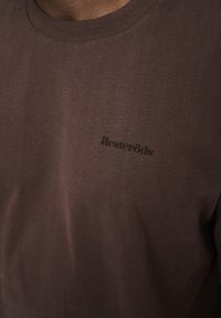 Brun t-shirt lavet af blød bomuld, med rund halsudskæring og et lille, mørkt trykt logo med teksten "Resteröds" på brystet.