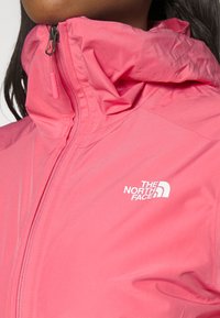 Chaqueta impermeable rosa con un alto cuello, con una textura suave y un logo blanco en el lado izquierdo. La cremallera está parcialmente abierta.