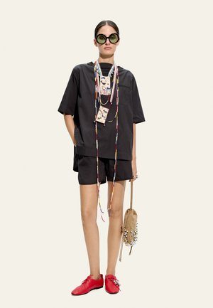 Kobieta nosząca oversize czarną koszulę i szorty, duże okrągłe okulary przeciwsłoneczne, kolorowe naszyjniki z koralików, czerwone buty i trzymająca beżową torebkę z koralikami.