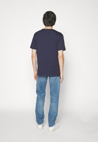 Μπλε βαθυά βαμβακερή μπλούζα T-shirt με κοντά μανίκια, κανονική εφαρμογή, στρογγυλή λαιμόκοψη, εμφανίζεται από πίσω, συνδυασμένη με ανοιχτόχρωμα τζιν παντελόνια.
