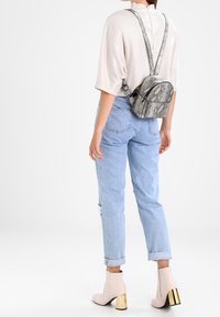 Mochila gris con estampado de piel de serpiente y correas ajustables, llevada sobre una blusa beige clara y unos vaqueros azul claro con dobladillos, combinada con botas de tacón beige.