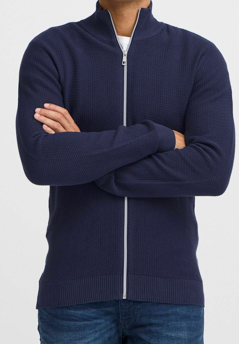 Homme portant un pull zippé bleu marine texturé avec un col montant et les bras croisés, associé à un jean bleu.