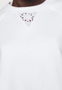 Vit sweatshirt med en triangulär utskärning vid halsringningen, prydd med delikat blommig broderi. Mjuk textur, rund krage och avslappnad stil.