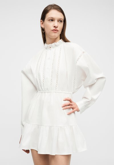 Γυναίκα με λευκό, μακρυμάνικο, μίνι φόρεμα με βολάν, λεπτομέρειες με κουμπιά και δαντέλα, ποζάροντας με το ένα χέρι στη μέση.