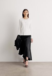 Weißes Langarmoberteil, schwarzer, fließender Rock mit welligem Saum, schwarze, rückenfreie High Heels und eine schwarze Jacke über dem Arm, minimalistische Umgebung.
