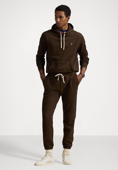 Temno rjav hoodie s kengursko žepom in vrvico, v kombinaciji s usklajenimi trenirkami z elastičnimi manšeti, obut z lahkimi supergami.