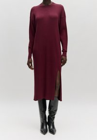 Robe en tricot côtelé bordeaux à manches longues, avec fente latérale et coupe décontractée. Associée à des bottes noires ajustées jusqu'au genou.