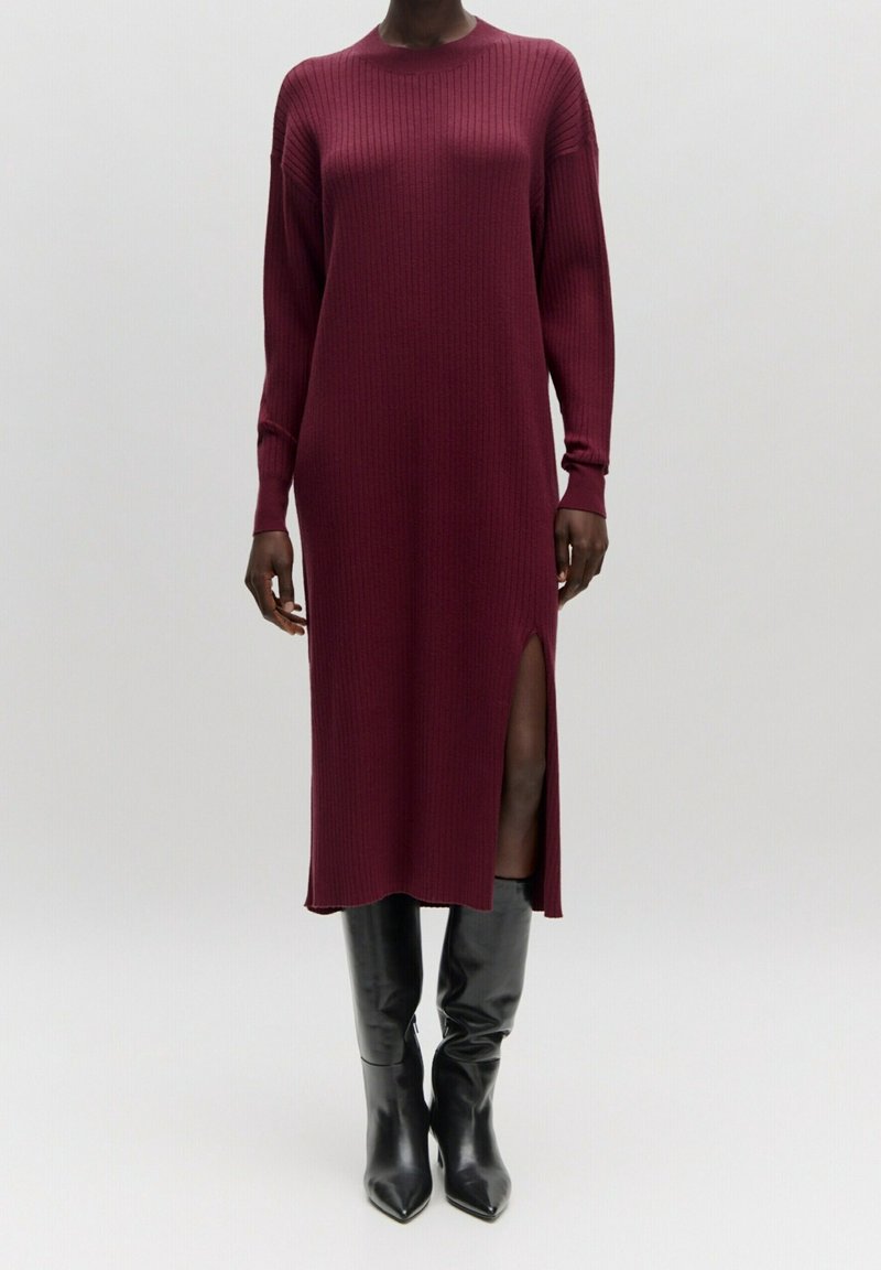 Robe en tricot côtelé bordeaux à manches longues, avec fente latérale et coupe décontractée. Associée à des bottes noires ajustées jusqu'au genou.