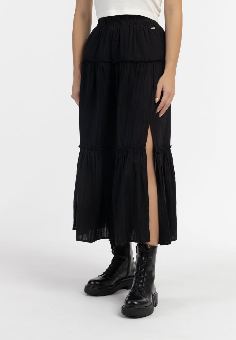 Jupe longue noire à volants avec une taille élastique froncée, une fente latérale et un tissu texturé ; associée à des bottes noires à lacets.