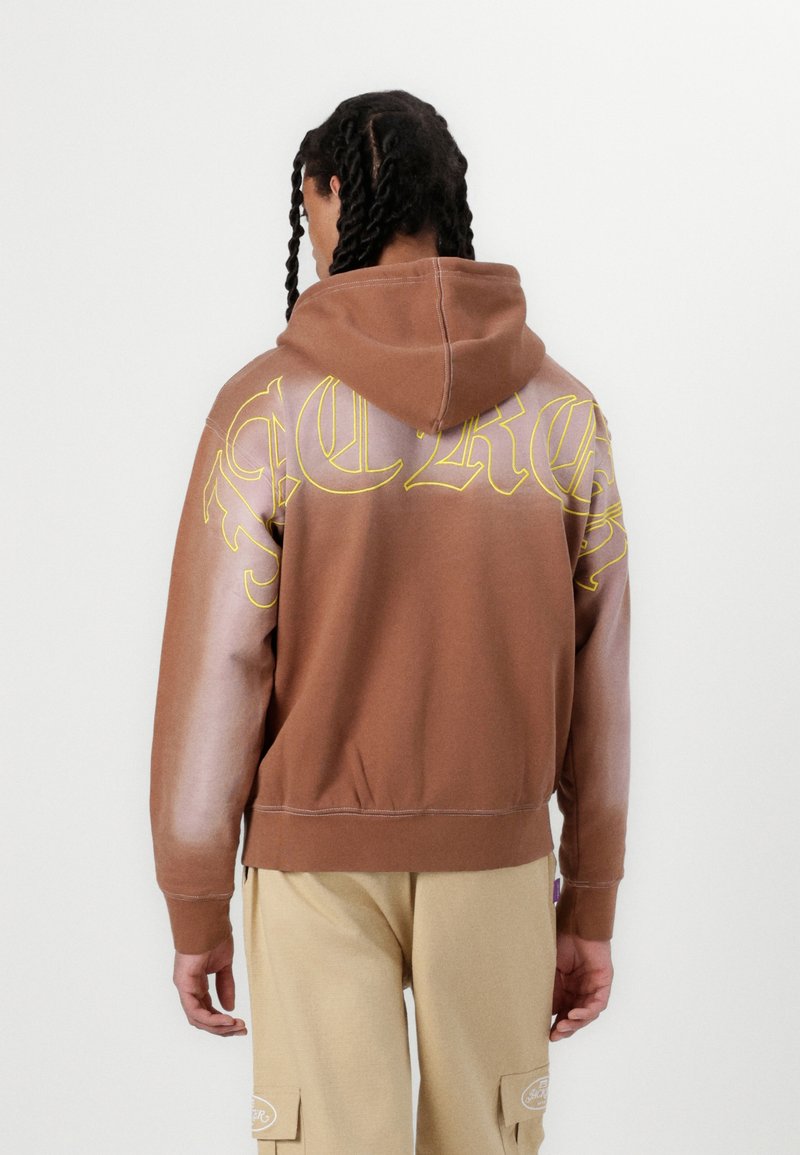 Brown hooded sweatshirt with yellow graphic text, lighter gradient sleeves, ribbed cuffs, and hem. Fabric appears soft and textured.