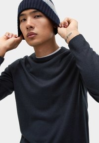 Dunkelblauer gestrickter Pullover mit Rippenbündchen. Getragen mit einer gestreiften Strickmütze. Verfügt über einen Rundhalsausschnitt und eine glatte Textur.