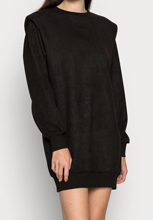 Vrouw draagt een zwarte sweatshirtjurk met lange mouwen, gewatteerde schouders en geribde boorden en zoom, staande tegen een effen achtergrond.
