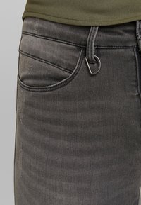 Pantalones vaqueros de mezclilla gris oscuro con detalles de bolsillo frontal, trabilla para cinturón y un anillo en D metálico plateado adjunto cerca de la cintura.