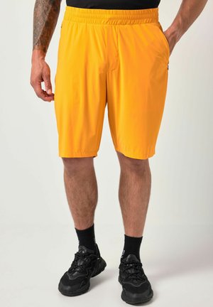 Mand iført lyse gule shorts, sort t-shirt, sorte crew-sokker og sorte sportssko, stående med den ene hånd i lommen.