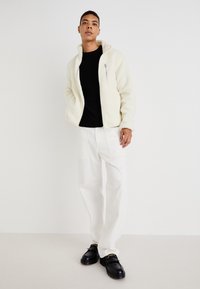 Casaco de fleece off-white com um bolso com zíper, combinado com um suéter preto em malha e calças brancas de perna larga. Sapatos de couro pretos com acabamentos dourados.