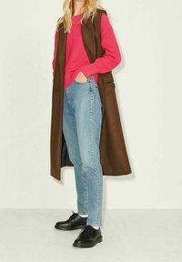 Gilet lungo marrone sopra un maglione rosa a costine, abbinato a jeans azzurri chiari e scarpe nere. Tessuto liscio con hardware minimal.