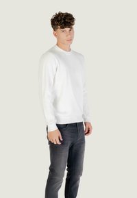 Witte geribbelde gebreide trui met lange mouwen en ronde hals, gecombineerd met donkergrijze jeans. Model staat voor een neutrale achtergrond.