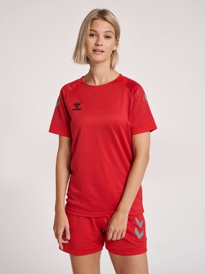 Vrouw draagt een rood sportshirt en bijpassende shorts met een grijs Chevron-logo, staand tegen een effen lichte achtergrond.