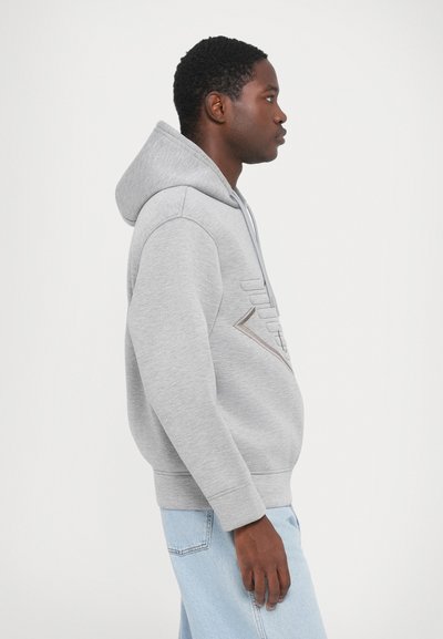 Grijze hoodie gemaakt van zachte stof, met een logo-design op de voorkant, een trekkoordkap en ribboorden. Gekocht met lichtblauwe jeans.