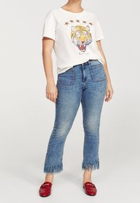 T-shirt bianco con grafica di una tigre e cinque stelle, abbinato a jeans blu con orlo sfrangiato e scarpe rosse slip-on.