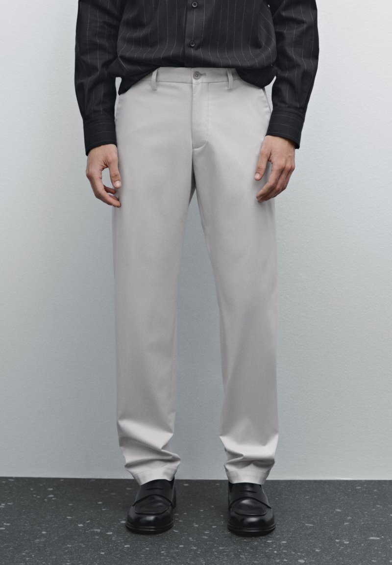 Beige Anzughosen mit einer glatten Textur, tailliertem Schnitt, seitlichen Taschen und geradem Bein, kombiniert mit schwarzen Dressschuhen.