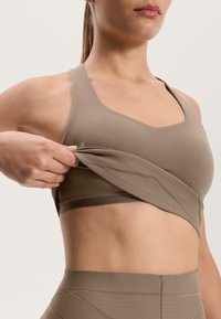 Béžový športový podprsenka s výstrihom do V, pružným materiálom a textúrovaným pásom. Nosička si upravuje látku, aby odhalila vrstvený vzhľad.