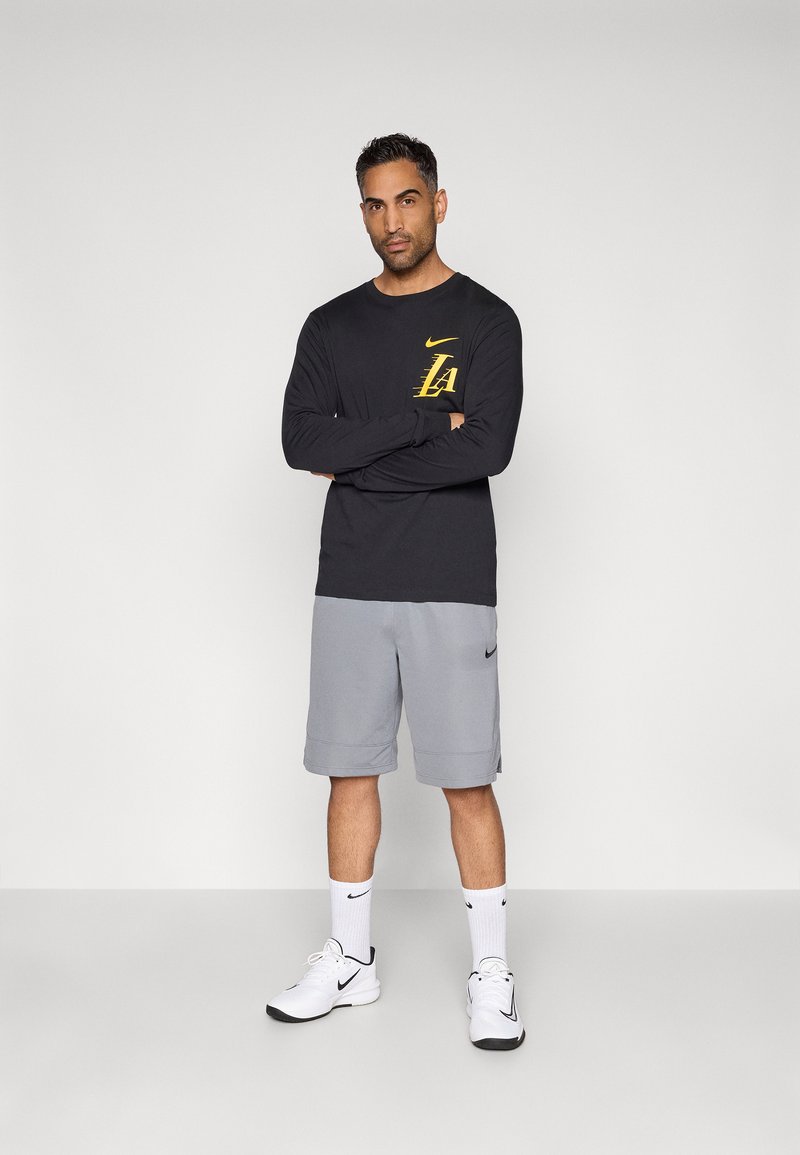 Camisa negra de manga larga con el logo amarillo de Nike, combinada con pantalones cortos grises. Zapatillas atléticas blancas y calcetines completan el atuendo deportivo.
