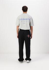 Gray short-sleeve T-shirt with "ALLSAINTS" printed in purple on the back. Black pants with a graphic detail and two back pockets. White sneakers.