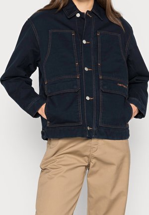 Henkilö pukeutuneena tummansiniseen napilliseen työjakkiin, jossa on suuret etutaskut, ja vaaleanruskeisiin housuihin, kädet takataskuissa.