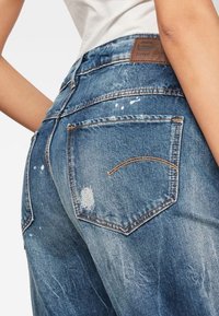 Denim jeans featuring a faded blue color, distressed texture, and back pockets with stitching details, including a brown leather patch.