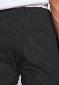 Gros plan d'un pantalon noir à taille élastique avec une unique poche passepoilée à l'arrière, porté avec une chemise blanche.