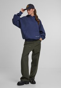 Marineblauwe hoodie met een voorzak, gecombineerd met donkergroene wijde broek. Het model draagt een zwarte pet met een rond logo en stevige zwarte schoenen.