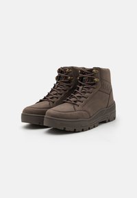 Botas de tobillo marrones con cordones, suelas gruesas, ojales de metal y cuellos acolchados, ubicadas sobre un fondo blanco.