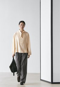 Vrouw loopt binnen, draagt een beige blouse, losse grijze broek, donkere schoenen en houdt een zwarte tas in haar linkerhand.