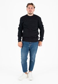 Giovane in piedi, indossando un felpa nera, jeans blu e sneakers bianche su uno sfondo bianco semplice.