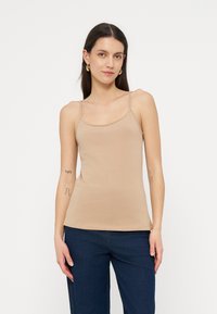 Kvinne med langt mørkt hår som har på seg gullfargede creol-øredobber, beige ermeløs camisole og mørkeblå bukser, stående foran en hvit bakgrunn.