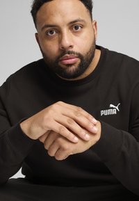 Felpa nera a girocollo in cotone misto, con logo bianco Puma sul lato sinistro del petto. Le mani del modello sono incrociate davanti.