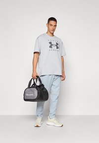 Grå oversized t-skjorte med "Under Armour" logo, lyseblå joggebukser og kremfargede sneakers. Svart og grå treningsbag med "Champion" logo.
