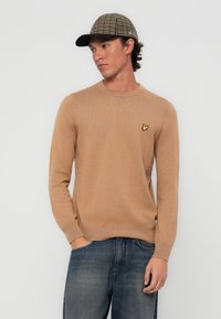 Beige pulóver kerek nyakkal, bordás mandzsettákkal és egy kis sárga hímzett logóval. Kék farmernadrággal és kockás sapkával párosítva.