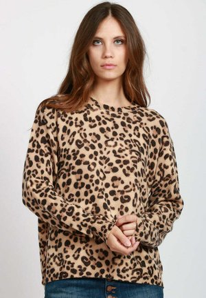 Donna con lunghi capelli castani, che indossa un maglione beige con stampa leopardata e jeans blu, in piedi di fronte a uno sfondo chiaro e semplice.