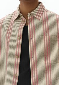 Camisa de botones a rayas con finas líneas beige, marrón y rosa, con un bolsillo en el pecho y cuello redondeado; hecha de tela ligera.